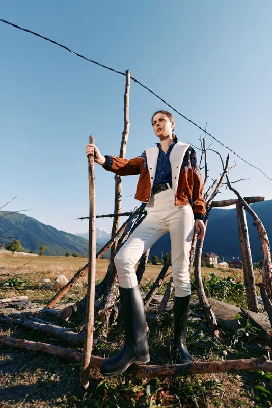 Woman in countryside standing on a wooden fence holding a walking stick, wearing shearling jacket, pants and rubber boots, with mountains and clear sky in the background.
