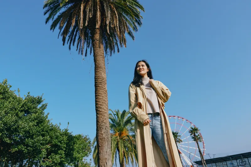 Woman in a long coat outdoors among palm trees with a ferris wheel in the background, fashion street style, sunny day, confident pose and lifestyle vibe