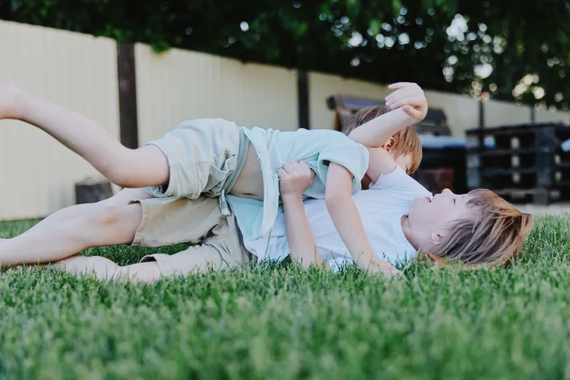 Two playful boys wrestle on a lush green lawn, showcasing joyful moments of childhood and camaraderie, radiating fun and energy in a sunny setting.