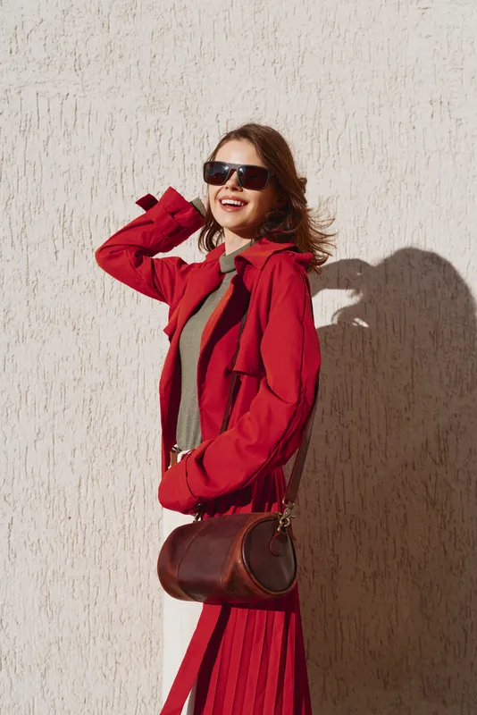 Young woman in a red coat and sunglasses smiles confidently while standing against a textured wall in bright sunlight during daytime.