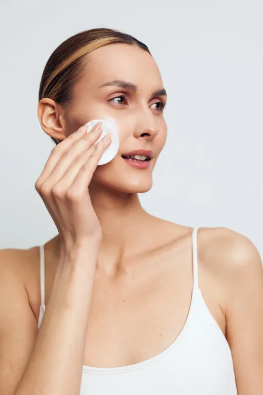 Woman skincare beauty face hygiene cleansing, cotton pad touches cheek, calm expression, close up portrait on bright studio background, self care routine for skin health