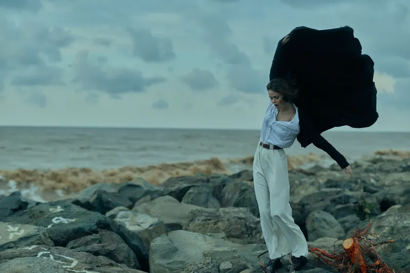 Wind sweeps along the coast as a woman in white flows with a dramatic cape, capturing motion against a rocky shore and vast seascape.