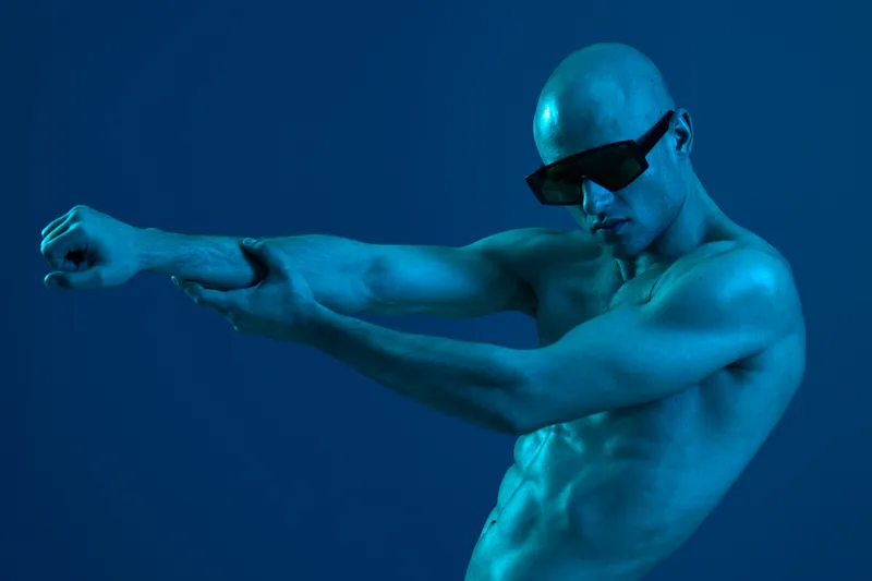Athletic man wearing sunglasses stretching under dramatic blue lighting, showcasing strength and confidence in a striking pose. Perfect for health and fitness themes.