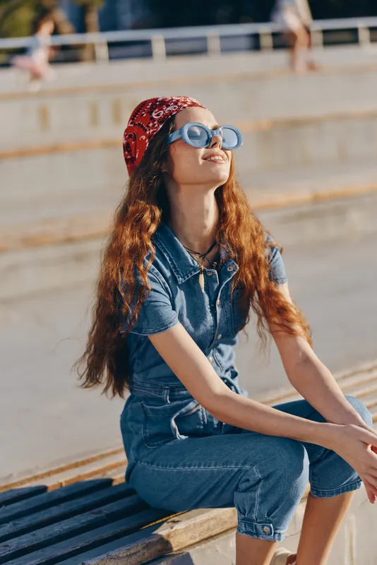 lifestyle woman wearing modern boho-western denim outfit with sunglasses and red bandana sitting outdoors, film color street style capturing warm sunlight and relaxation