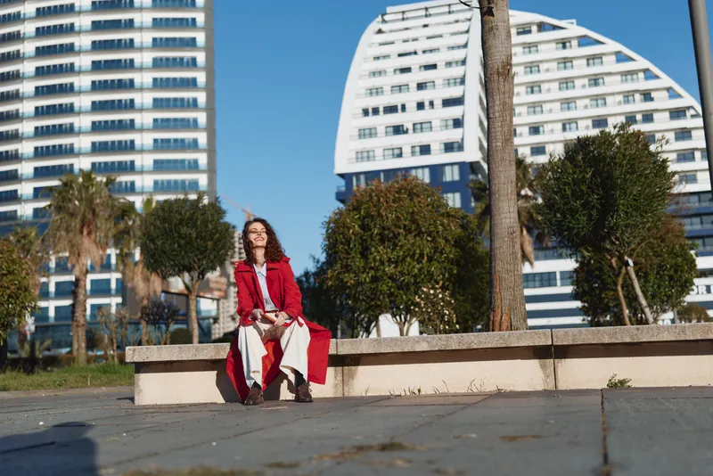 Young woman in red coat sits on concrete bench in urban park with modern buildings background under sunny clear sky, enjoying peaceful moment outdoors.