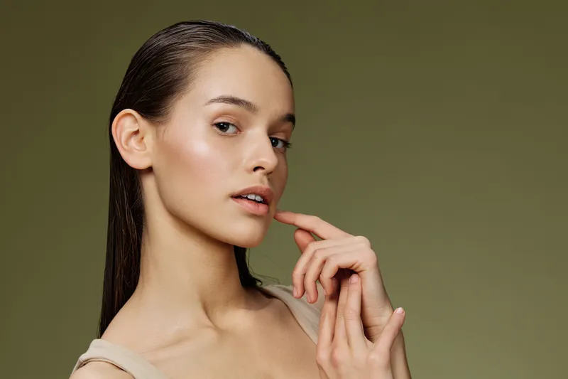 A close up portrait of a young model with straight hair, wearing a neutral colored top, showcasing a calm expression against a soft green background, emphasizing natural beauty and healthy skin