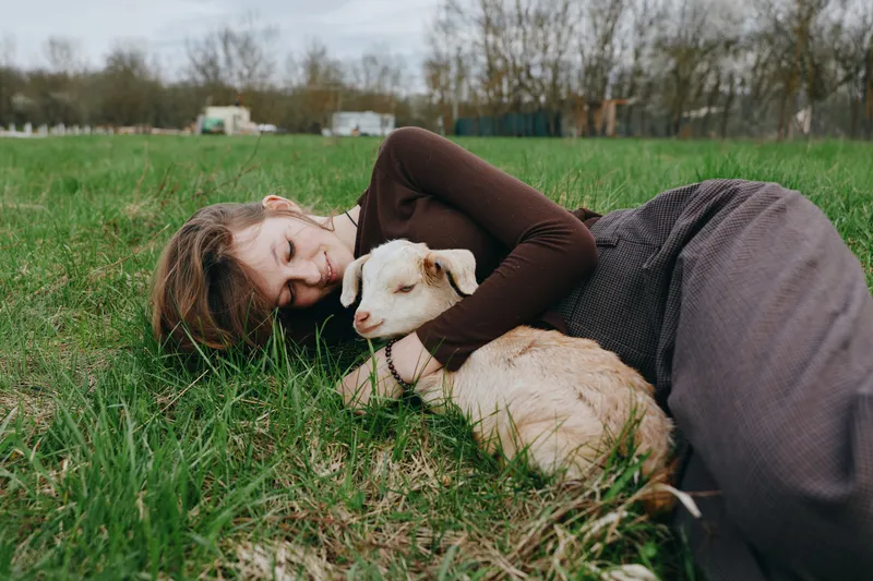 Warm outdoor moment with a woman lying in fresh grass, hugging a gentle goat kid, soft earth tones, relaxed mood, sunlit snuggle, rural lifestyle, carefree connection with animals