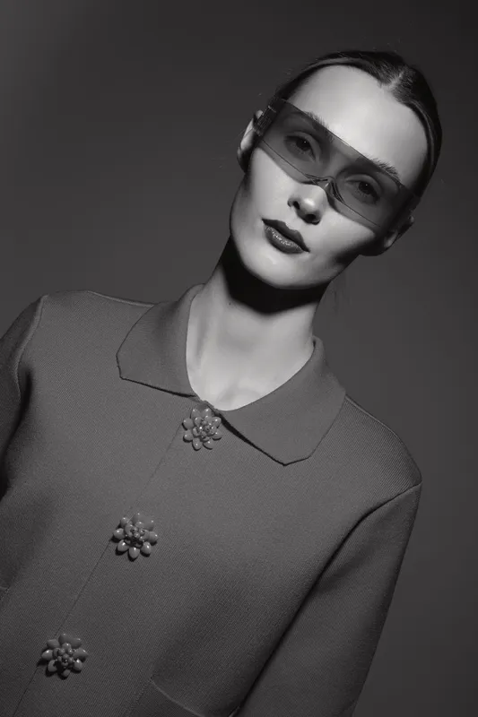 Young Caucasian woman wears stylish sunglasses and textured coat with decorative buttons, posing in monochrome studio lighting.