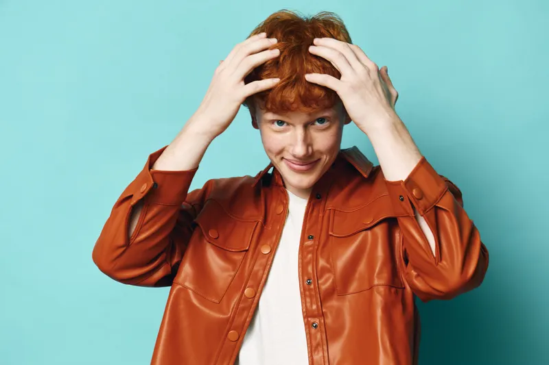 Redhead man with curly hair and smiling face in brown leather jacket on blue background. Hands touching hair, casual style, confident expression and relaxed pose in studio.