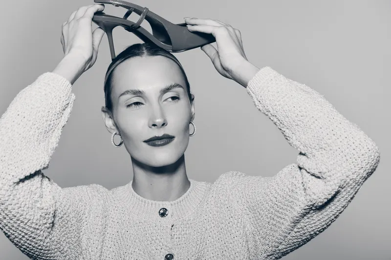 Young white woman with pale skin and slicked back hair wearing a textured sweater putting on sunglasses. Studio portrait with minimalist lighting and neutral background. Fashion concept.