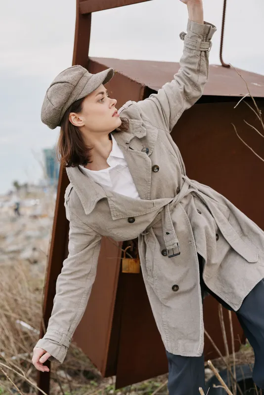 Young woman in beige trench coat and hat poses outdoors near rusty metal structure. Autumn fashion style with neutral tones and casual mood in natural environment.