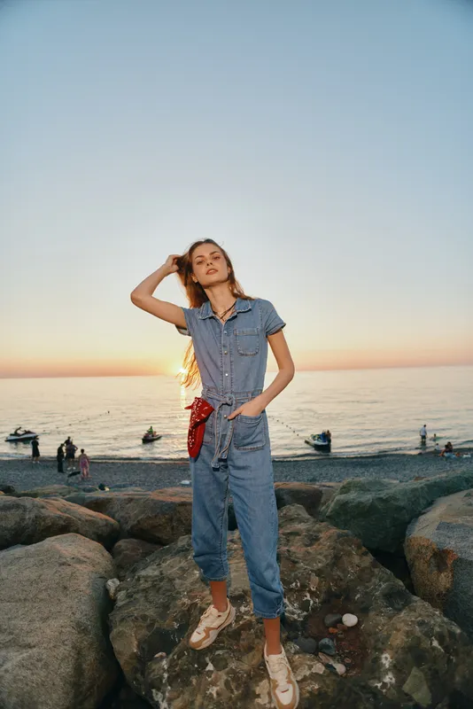 Lifestyle woman in modern boho-western denim street style stands confidently on rocks by the sea during sunset, captured with warm film color tones.