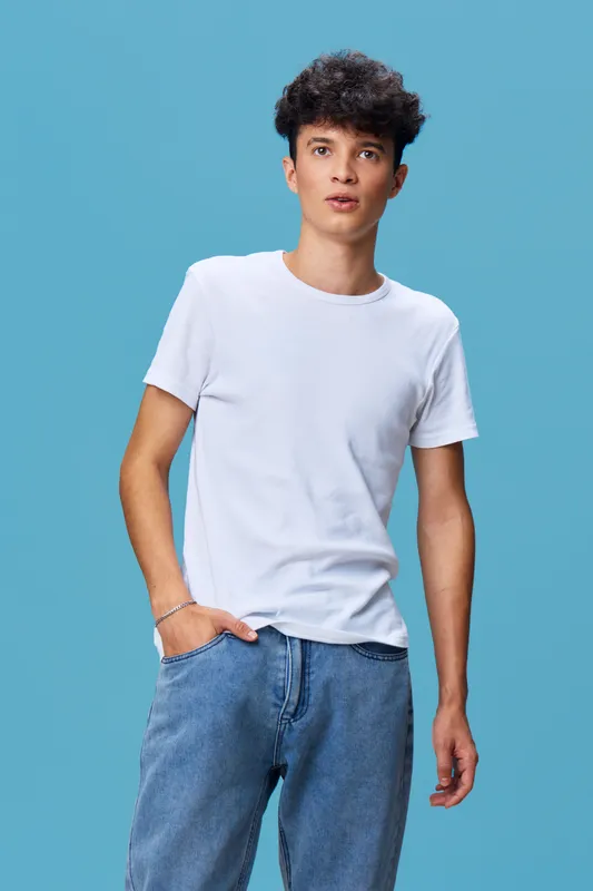 young man in white t-shirt and jeans standing confidently against blue background with casual posture and curly hair, natural light enhances relaxed mood