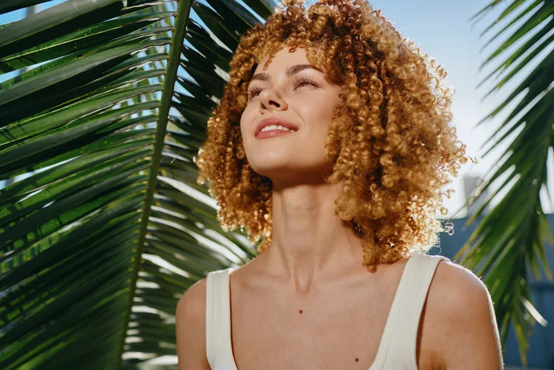 smiling woman with curly hair enjoying sunshine outdoors near green tropical palm leaves on a bright summer day in casual white top