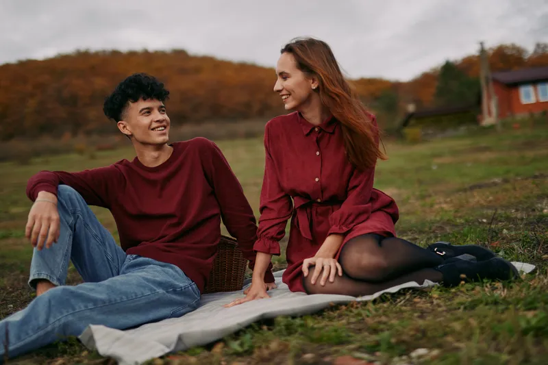 Young couple sharing a genuine outdoor moment on a picnic blanket in an autumn meadow, relaxed smiles and natural gestures conveying authentic connection and everyday intimacy.