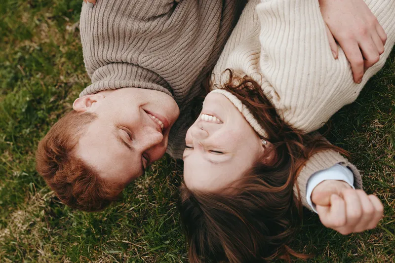 Couple outdoors laughter smiles, lying on grass in soft sweaters, capturing affection and a sunlit, relaxed moment, natural colors and carefree mood, lifestyle portrait focusing on connection and