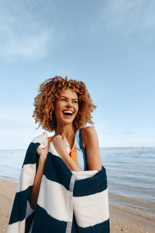 A young woman with curly hair smiles on a sunny beach, wrapped in a striped towel, conveying joy, warmth, and carefree summer vibes.