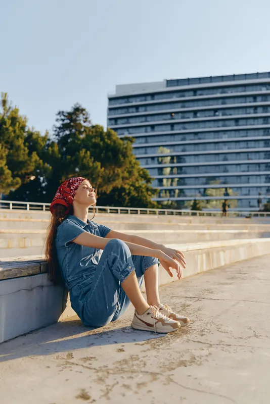 Lifestyle woman in modern boho-western denim street style sitting relaxed outdoors enjoying sunny day with film color aesthetic and urban background.