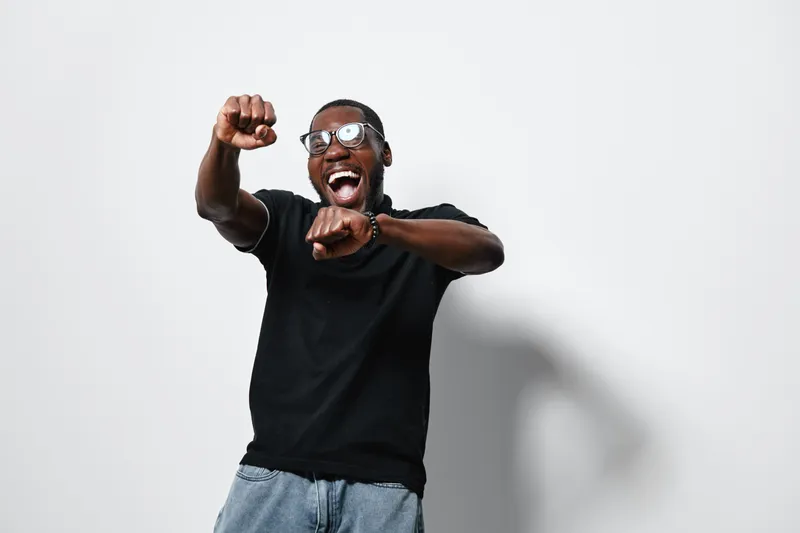Black man with dark skin wearing glasses and black t shirt dancing and smiling with excitement against white background. Happy African American male expressing joy and energy.