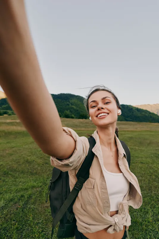 Smiling woman with backpack taking a selfie in a green meadow. Outdoor lifestyle, adventure, and wellness vibe under soft daylight, showcasing confidence, freedom, and connection with nature.