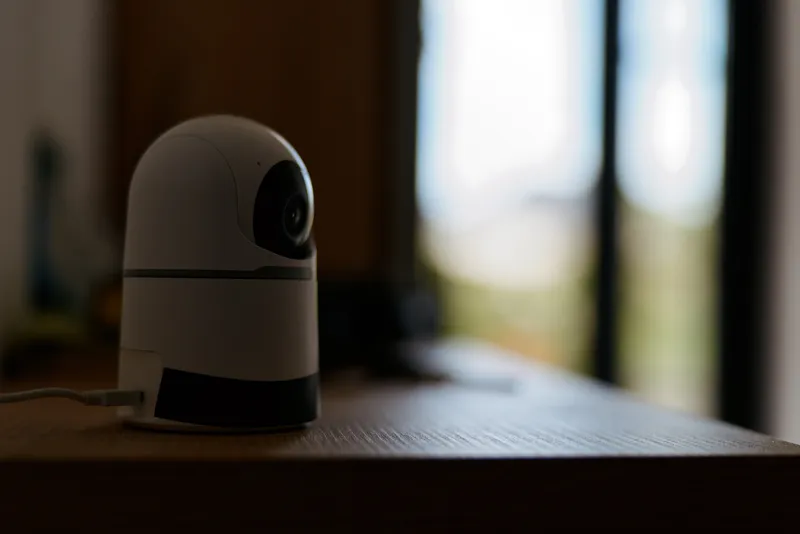 A small security camera sits on a wooden desk near a bright window, capturing a calm home scene with a minimal, modern touch.