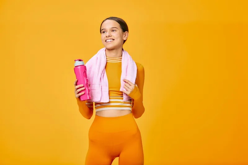 Woman fitness, sport enthusiast standing in a vibrant orange studio with a pink towel around her neck and a pink water bottle after a workout, athletic and confident, full-body shot, energy and
