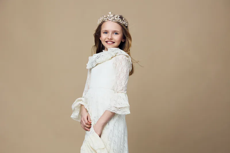 Young girl in a delicate white dress with a crown, smiling joyfully against a neutral beige background, embodying innocence and elegance in a playful, whimsical style