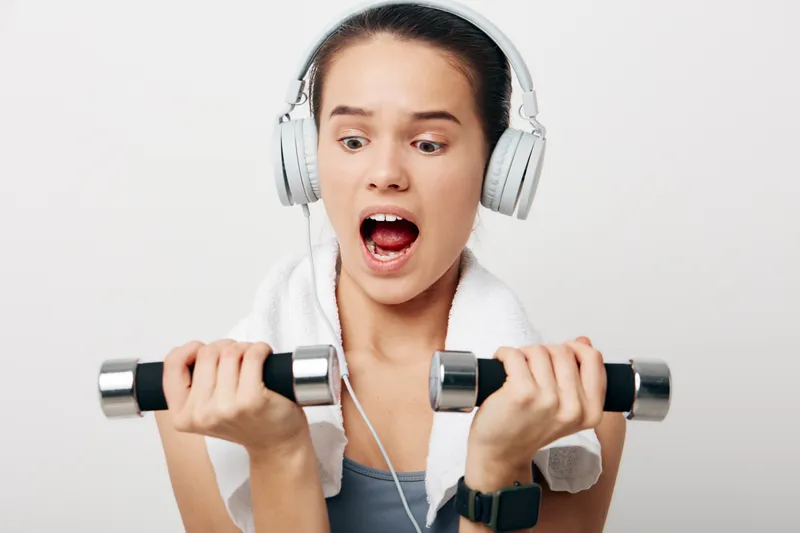 Young woman fitness portrait lifting chrome dumbbells while wearing headphones and towel, surprised expression and focused effort, indoor studio with clean white background, sport concept.