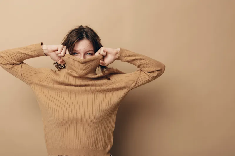 Young woman in beige sweater pulls collar over face, peeking with eyes. Neutral background and casual style create cozy, playful mood. Concept for autumn, warmth, and comfort.
