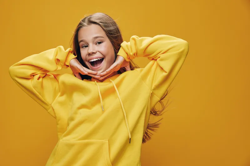 Girl smiling and posing with hands under chin wearing yellow hoodie with long hair on yellow background. Young female happy and cheerful with casual outfit and bright mood.