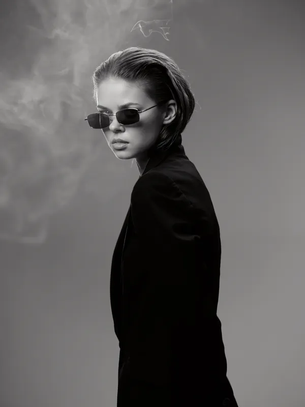 Black and white portrait of stylish woman wearing sunglasses and black blazer with smoky background, exuding confidence and modern elegance.