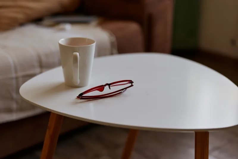 A minimalist roundtable holds a white mug and red framed glasses on a smooth surface, while a cozy living room blur creates a warm, intimate home atmosphere.