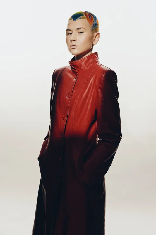 woman with colorful short hair wearing a red leather coat, standing with hands in pockets against plain light background, modern high fashion style
