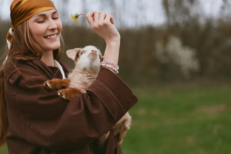 A smiling woman in a brown cloak gently admires a goat kid, lifting it toward a fluttering butterfly in a sunny field. A lifestyle moment focusing on animal care, curiosity, and simple countryside