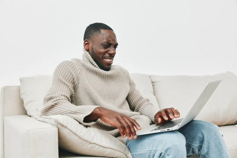 African man in cozy sweater looking frustrated at laptop, sitting on light colored sofa, expressing emotions of stress and discomfort while working from home