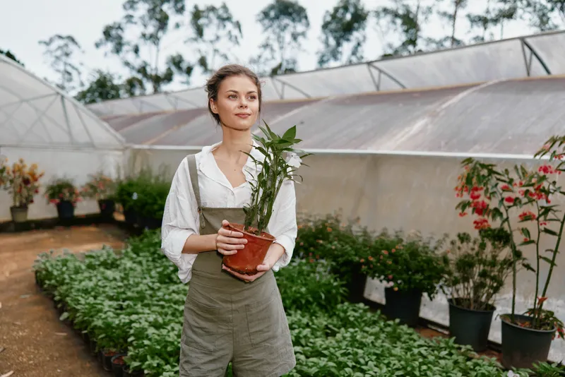 Young woman wearing an apron in a greenhouse, holding a potted plant, surrounded by greenery and flowers, representing gardening and healthy living