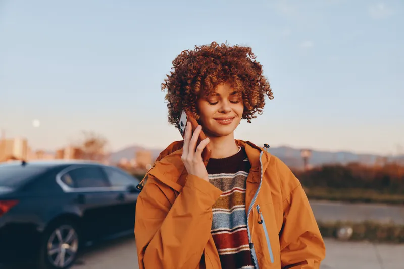 Woman lifestyle outdoors, smiling with curly hair, wearing a warm jacket, standing on a street near a parked car, soft sunset light, relaxed expression, urban blur background