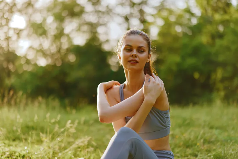 Young woman in activewear enjoying a moment of reflection in nature, expressing tranquility and well being, surrounded by green foliage in soft sunlight