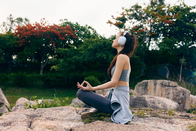 Young woman meditating outdoors in stylish sportswear, headphones on, enjoying tranquility in a serene green park, surrounded by fall foliage