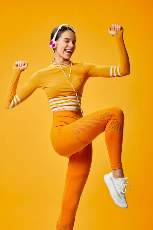 Vibrant orange studio scene showing a smiling woman in athleticwear performing a dynamic fitness pose with headphones, celebrating movement, energy and sport performance.