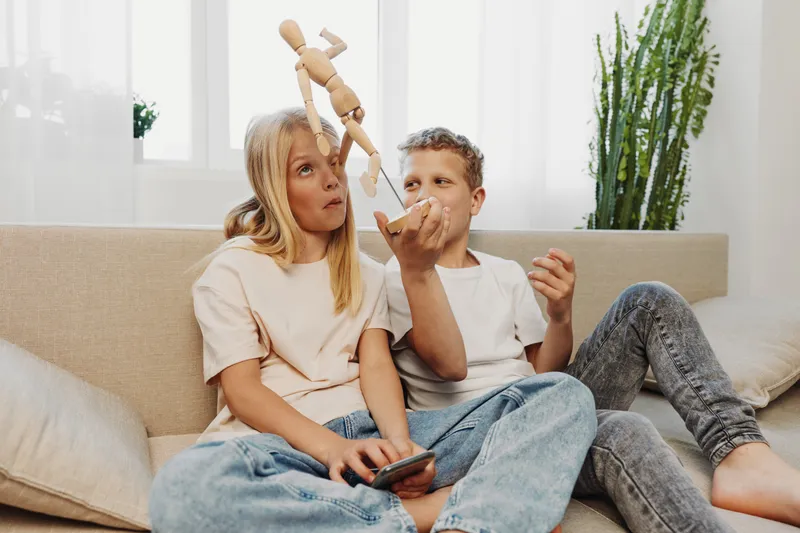 Curious children sitting on couch playing with wooden mannequin and remote control, bright living room interior with plants and natural light.