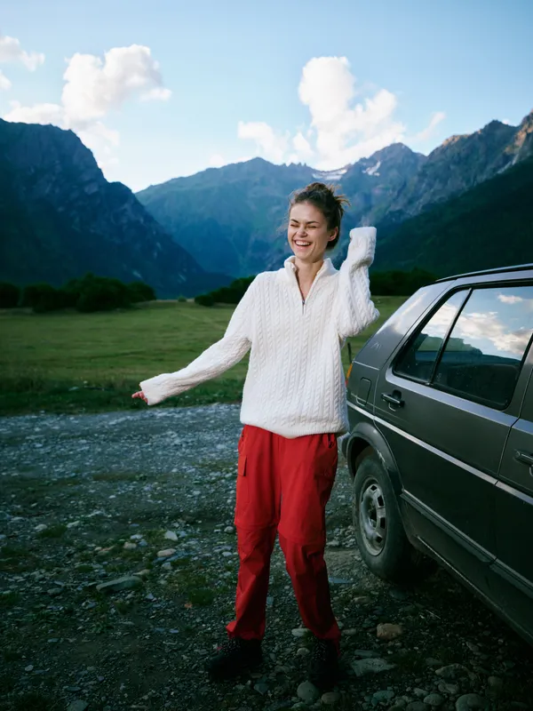 Outdoor woman stands beside a car in a mountainous landscape, wearing a white sweater and red pants, feeling free, joyful, and energized as sunlit peaks rise behind.