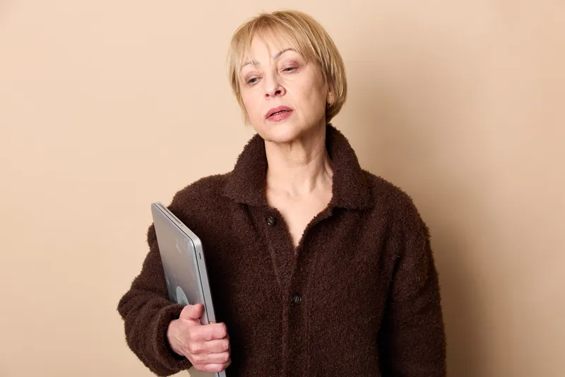 Woman holding tablet in studio beige background, mature blonde short hair, thinking neutral expression, portrait framing, communication with digital technology, soft even light, indoor minimal setting