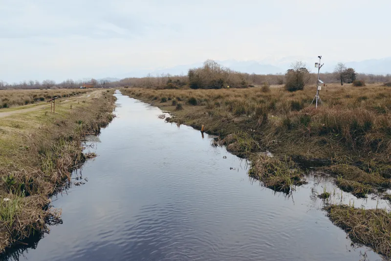 River canal in a rural landscape with calm water reflecting cloudy sky, natural grassy banks and shrubs, outdoors nature scenery, peaceful lifestyle mood, wide view, long scenery