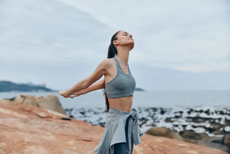 Young female model practicing yoga on a rocky beach, embodying calmness and serenity in a gray sport outfit against a serene ocean backdrop