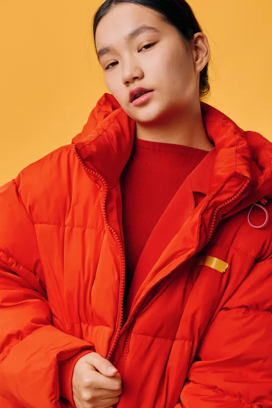 Young woman in red puffer jacket posing against orange background, fashion portrait with casual style and confident expression for winter clothing or seasonal outfit.
