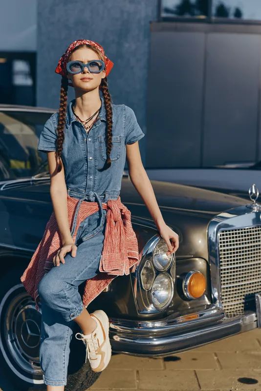 young woman with braids wearing sunglasses and denim outfit leaning on vintage car in urban setting with casual style and bright sunlight