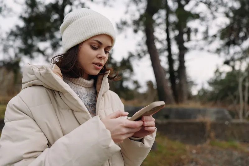 Woman using smartphone outdoors in winter coat and beanie, focused on the device in a park with trees, calm atmosphere, casual urban lifestyle