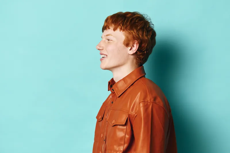 Profile of young man smiling and looking forward on blue background wearing brown leather shirt with casual hairstyle and positive expression for lifestyle portrait.