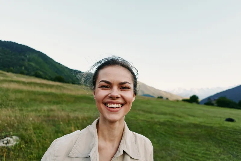 Smiling woman in an outdoor portrait in a meadow with distant mountains, wearing a beige jacket. Casual lifestyle and nature vibe with warm daylight.